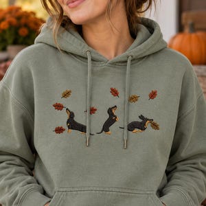 Könnte beinhalten: Ein olivgrüner Kapuzenpullover mit einer Stickerei von drei schwarz-braunen Dackeln, die mit verstreuten Herbstblättern in Orange-, Gelb- und Brauntönen spielen. Der Hoodie verfügt über eine Kängurutasche auf der Vorderseite und Kordelzüge.