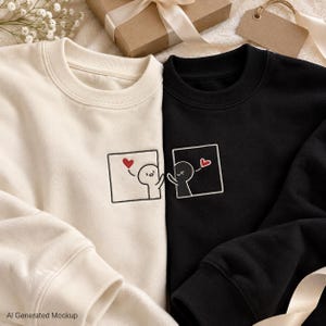 Könnte beinhalten: Ein cremefarbenes Sweatshirt und ein schwarzes Sweatshirt, jeweils mit einem gestickten quadratischen Rahmen, der eine einfache Cartoon-Figur und ein kleines rotes Herz enthält. Die Figuren sind so platziert, als würden sie sich über die Kleidung hinweg erreichen.
