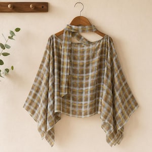 May include: A beige and gold plaid patterned top with a neck tie, hanging on a wooden hanger. The top has a cape-like design with wide sleeves. The fabric appears lightweight and airy, suitable for warmer weather.
