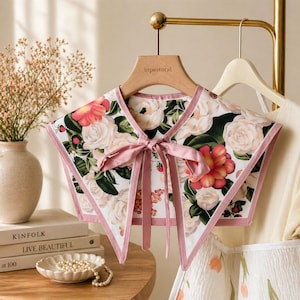 May include: A floral collar with a white background, featuring pink and red roses and green leaves. The collar has a pink ribbon tie and a pink border. The brand name "inpastoral" is visible on the wooden hanger.