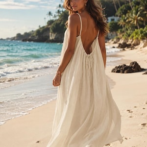 Puede incluir: Un vestido largo blanco roto con tirantes finos y espalda baja. El vestido está hecho de una tela ligera y se lleva en una playa de arena con el mar de fondo. Ideal para unas vacaciones en la playa.