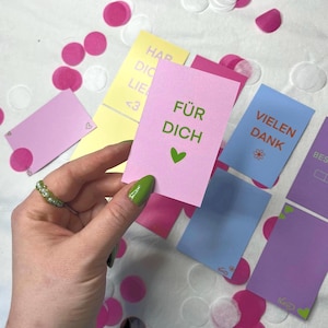 Könnte beinhalten: Eine rechteckige rosa Karte mit dem grünen Text FÜR DICH und einem grünen Herzsymbol. Weitere bunte rechteckige Karten liegen verstreut daneben, mit verschiedenen deutschen Phrasen wie VIELEN DANK und HAR DIC LIEB, sowie rosa und weißem Konfetti.