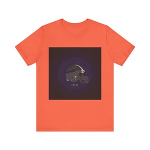 Pode incluir: Camiseta laranja com um gráfico de um capacete de futebol americano. O capacete é cinza e dourado, em um quadrado roxo escuro com círculos concêntricos. A camiseta é feita de um material macio e confortável.