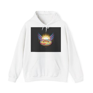 Puede incluir: Sudadera con capucha blanca con un gráfico en la parte delantera. El diseño presenta un sol con alas y un círculo interior de colores del arcoíris, sobre un fondo negro. La sudadera tiene un bolsillo delantero y una capucha con cordón.