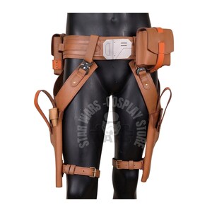 May include: A brown leather utility belt and holster set. The belt features a silver buckle and multiple pouches with orange accents. Holsters are attached to the legs with straps. The set is displayed on a black mannequin.
