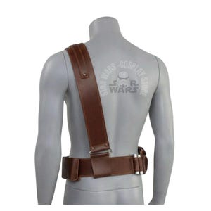 May include: A brown leather shoulder strap and belt set, likely for a costume. The strap is wide and attaches to the belt with metal hardware. The belt has multiple pouches and silver-colored accents. The mannequin has a Star Wars logo on the back.