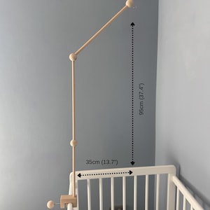 Op de afbeelding: Een lichtgekleurde houten mobielarm met een klemvoet, verticale en schuine stangen verbonden door bolvormige gewrichten, en een haak aan het uiteinde. De arm meet 95 cm in hoogte en 35 cm in breedte en is bevestigd aan een wit ledikant.