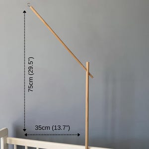 May include: A light-colored wooden mobile arm stand featuring a vertical post, a diagonal extension arm with a metal hook at the top, and a clamp base. The stand measures 29.5 inches in height and 13.7 inches in width, attached to a white crib rail.