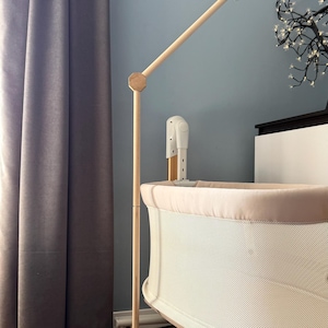 May include: A light-colored wooden mobile stand with a tall vertical pole and an angled arm, positioned next to a beige and white mesh bassinet. The stand features a hexagonal joint connecting the two wooden segments, providing a sturdy structure.
