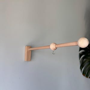 Könnte beinhalten: Eine hellfarbene Wandhalterung aus Holz mit rechteckiger Basis, zylindrischem Arm und kugelförmigem Endstück. Eine verschiebbare Holzkugel mit einem kleinen silberfarbenen Metallhaken an der Unterseite ist auf der horizontalen Stange angebracht.