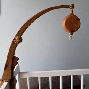 May include: Wooden baby mobile with a curved arm and a round, bell-shaped element. The mobile is made of light-colored wood and is attached to a white crib. The design is simple and elegant, perfect for a nursery.