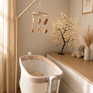 May include: A baby bassinet with light beige fabric sides and a wooden frame, accompanied by a hanging mobile featuring felt butterflies in shades of terracotta and cream, suspended from a tall wooden stand positioned next to the bassinet.