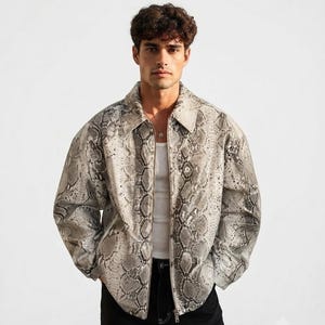 May include: A long-sleeve jacket featuring a grey and black snakeskin pattern, a pointed collar, and a full-length front zip closure. The jacket is worn over a plain white vest top and paired with black denim jeans with visible white stitching.