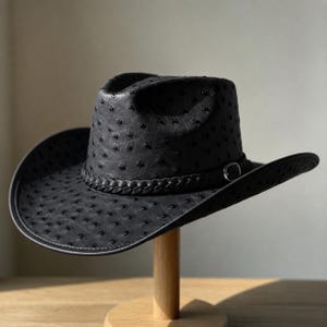 May include: A black cowboy hat featuring a textured ostrich-style finish with raised quill bumps. The hat includes a braided leather band around the crown, accented by a small silver-toned metal buckle on the side, and a wide, curved brim.