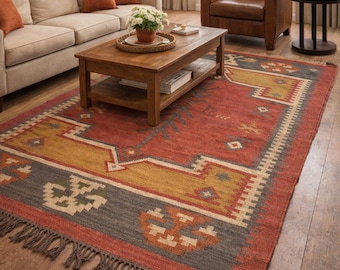 Puede incluir: Una alfombra rectangular con un patrón geométrico en tonos rojos, amarillos, grises y beige. La alfombra tiene un borde con flecos y está colocada sobre un suelo de madera en un salón. Se ve un sofá y un sillón.
