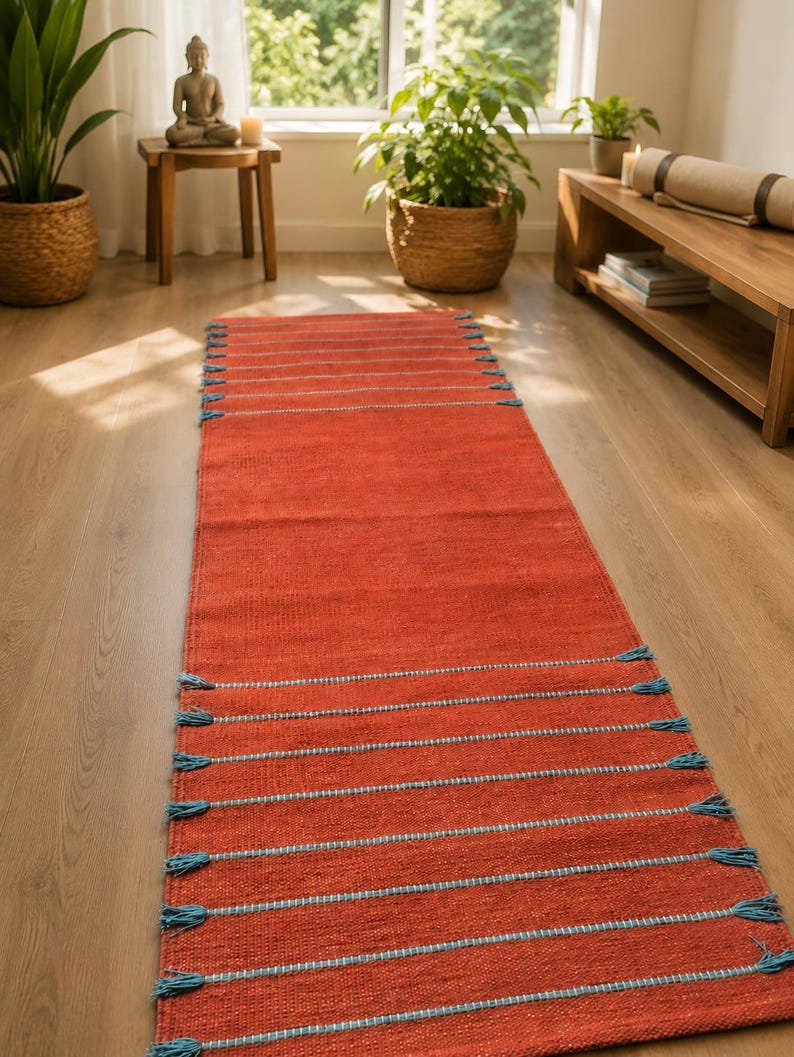 May include: A long rectangular runner rug in a vibrant terracotta orange color. The rug features horizontal stripes in a light blue hue, with small matching blue tassels attached at the ends of each stripe along both sides of the woven fabric.