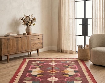 Puede incluir: Una alfombra rectangular con una base de color burdeos intenso y un patrón geométrico en tonos crema, rosa, naranja, verde azulado y gris oscuro. La alfombra tiene un borde ancho de color crema con un borde en zigzag y flecos cortos.