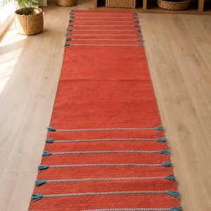 May include: A long, rectangular runner rug in a vibrant terracotta orange color. The rug features horizontal stripes in a contrasting light blue thread, with small, decorative blue tassels extending from the edges at each stripe interval.