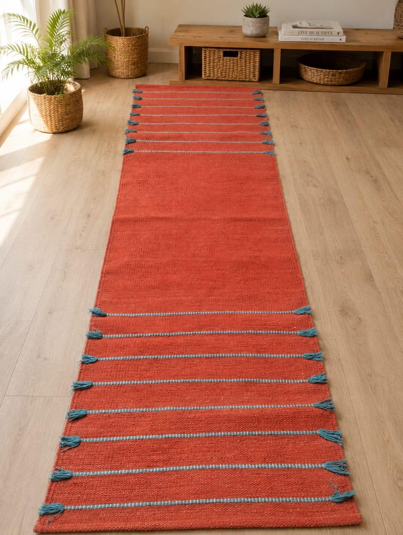 May include: A long, rectangular runner rug in a vibrant terracotta orange color. The rug features horizontal stripes in a contrasting light blue thread, with small, decorative blue tassels extending from the edges at each stripe interval.