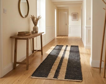 Puede incluir: Una alfombra de pasillo rectangular con textura tejida, que presenta franjas anchas negras alternadas con una franja central de color beige natural. La alfombra está colocada sobre un suelo de madera clara en un pasillo, extendiéndose a lo largo.
