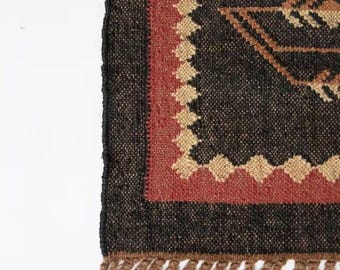Puede incluir: Una alfombra tejida con una base de color gris carbón oscuro, un borde rojo intenso y un patrón de diamantes de color tostado claro a lo largo del borde interior. La alfombra tiene una superficie texturizada y flecos de fibras marrones.