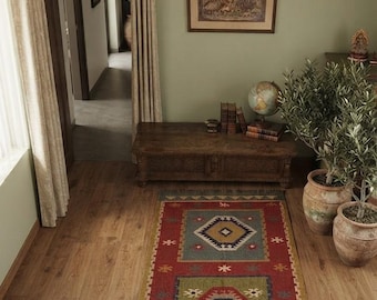 Puede incluir: Una alfombra rectangular tejida con fondo rojo, dos medallones geométricos centrales en azul y verde, pequeños motivos de estrellas y bordes con flecos. El texto "kartikrugs" aparece cerca del borde inferior de la alfombra.