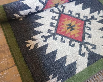 Puede incluir: Una alfombra tejida rectangular con un patrón geométrico en negro, blanco, rojo y amarillo mostaza. La alfombra tiene un borde verde oliva sólido y un fleco de borlas de color marrón claro en un lado. El texto KARTIKRUGS está impreso en negro.