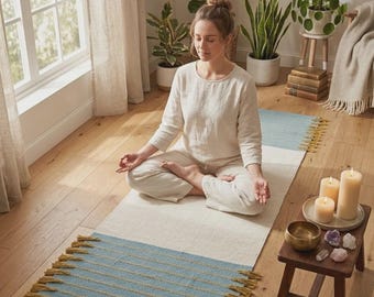 May include: A light blue and cream-colored yoga mat with gold tassels. The mat is rectangular and has a blue section with horizontal lines. A person is sitting on the mat in a meditative pose. Candles, crystals, and a singing bowl are on a small wooden table.