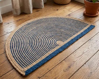Puede incluir: Una alfombra semicircular hecha de fibras de yute natural en tono bronceado, con patrones semicirculares concéntricos en azul marino profundo. La alfombra está terminada con un borde grueso trenzado en azul marino a lo largo del borde recto.