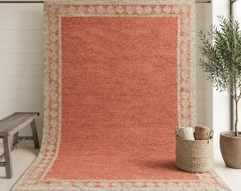 Puede incluir: Una alfombra rectangular grande con un centro de color naranja quemado sólido y un borde ancho con un patrón de diamantes repetitivo en crema y marrón claro. La alfombra tiene un tejido texturizado y flecos cortos en los bordes.