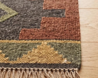 Puede incluir: Una alfombra tejida con una base gris carbón oscuro y patrones geométricos en naranja óxido, verde oliva y amarillo mostaza. La alfombra tiene un tejido grueso y texturizado, rematada con una hilera de flecos de color beige claro.