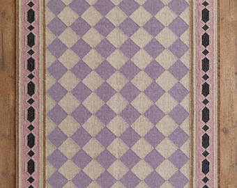 Puede incluir: Una alfombra rectangular con un patrón central de rombos en cuadrados alternos de color violeta claro y beige. La alfombra está enmarcada por un borde de formas geométricas negras sobre un fondo rosa, con flecos cortos en los extremos.