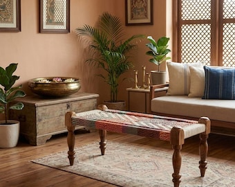 Puede incluir: Un banco rectangular de madera con cuatro patas torneadas y un asiento tejido con un patrón geométrico multicolor. El banco está colocado sobre una alfombra estampada en una habitación con paredes de color terracota, un baúl de madera y un sofá.
