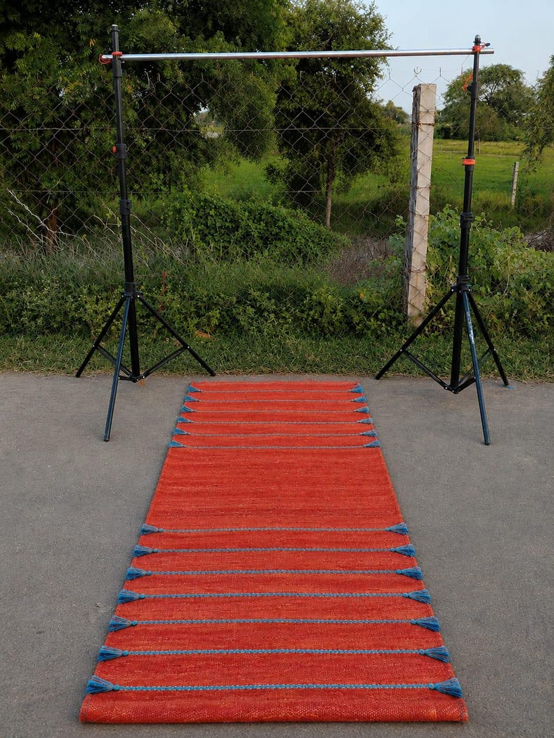 May include: A rectangular runner rug featuring a vibrant orange base color with horizontal rows of textured blue stitching. The rug is accented with small blue tassels attached at regular intervals along both long edges of the woven fabric.