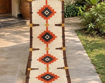 Puede incluir: Un camino de mesa largo y rectangular con una base de color crema y un patrón geométrico repetitivo. El diseño presenta motivos en forma de diamante en naranja, negro y amarillo mostaza, bordeados por bordes marrón oscuro con pequeños acentos de flecha.