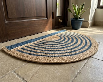 Puede incluir: Un felpudo semicircular hecho de fibras de yute natural trenzadas, con un patrón repetitivo de arcos concéntricos en azul y beige. La alfombra presenta un borde recto en la parte posterior y un borde frontal curvo con textura tejida.