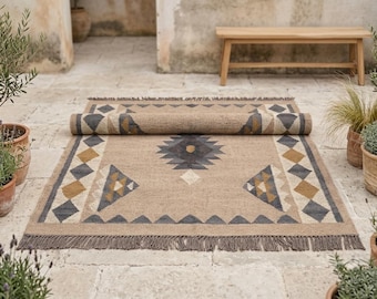 Puede incluir: Una alfombra rectangular con base color canela y un patrón geométrico en gris oscuro, amarillo mostaza y crema. La alfombra tiene bordes con flecos y está parcialmente enrollada en la parte superior, mostrando un diseño simétrico de rombos y triángulos.