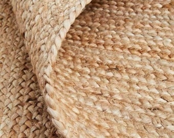 Puede incluir: Una alfombra tejida de color canela con un borde trenzado grueso y una superficie densa y texturizada hecha de fibras naturales. El material muestra un color marrón claro consistente con hebras entrelazadas que crean un patrón.