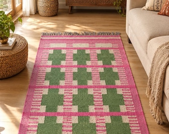 Puede incluir: Una alfombra rectangular con un patrón geométrico repetitivo en rosa vibrante, verde bosque y crema. El diseño consiste en formas de cruz verdes enmarcadas por rayas rosas y crema, terminada con flecos cortos de color gris en los extremos.