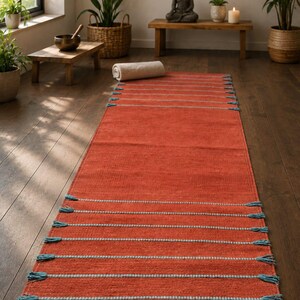 May include: A rectangular runner rug in a deep terracotta orange color featuring horizontal stripes of light blue thread. The rug is finished with small, matching light blue tassels at the ends of each horizontal stripe along both sides.