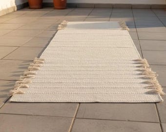 May include: A rectangular, off-white woven rug with a textured surface and fringe detailing on both ends. The rug is placed on a tiled surface, and the neutral color palette suggests a minimalist or bohemian style. The rug's dimensions are not visible.