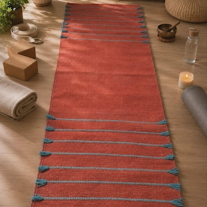 May include: A rectangular yoga mat in a deep terracotta orange color featuring horizontal stripes in a contrasting light blue. The mat is finished with small, decorative light blue tassels attached at the ends of each horizontal stripe along the sides.