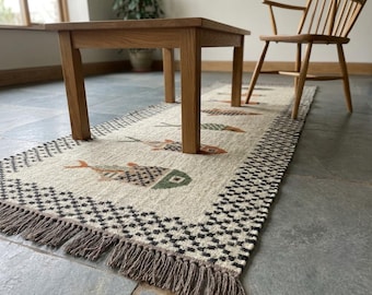 Puede incluir: Una alfombra de pasillo tejida rectangular con un patrón repetitivo de peces estilizados en tonos verde, naranja y marrón. La alfombra tiene un centro color crema, un borde a cuadros blanco y negro y flecos marrones en los extremos.