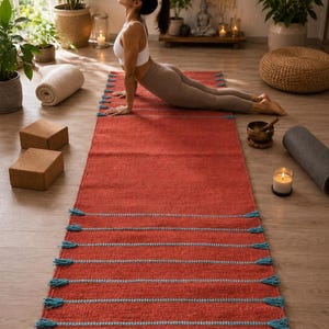 May include: A rectangular yoga mat in a deep terracotta red color featuring horizontal light blue woven stripes and decorative tassels along the edges. The mat has a textured surface and is positioned flat on a wooden floor surface.