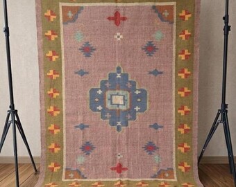 Puede incluir: Una alfombra tejida rectangular con un centro rosa polvoriento y un gran medallón geométrico azul. El borde es de color verde oliva con un patrón repetitivo de pequeños cuadrados amarillos y rojos. La alfombra tiene flecos en los extremos.