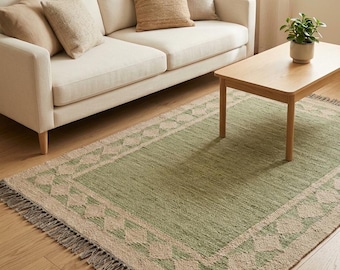 Puede incluir: Una alfombra rectangular con un panel central de color verde salvia sólido, enmarcado por un borde ancho con un patrón repetitivo de diamantes en color beige. La alfombra tiene flecos cortos y texturizados en los dos bordes más cortos.