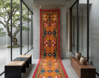 Puede incluir: Una alfombra pasillera rectangular tejida con una base de color naranja vibrante y patrones geométricos en rojo, verde, amarillo, azul y blanco. La alfombra cuelga de una barra horizontal de latón y se extiende hasta el suelo, donde está enrollada.