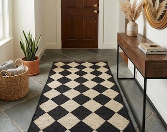 Puede incluir: Una alfombra rectangular con un patrón repetitivo de rombos en negro y beige. La alfombra tiene una textura tejida y está rematada con flecos cortos de color marrón oscuro en ambos extremos, creando un borde definido para el suelo.
