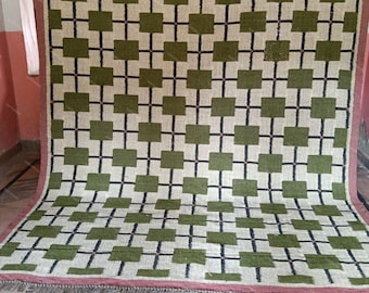 Puede incluir: Una alfombra rectangular con un patrón de cuadrícula repetitivo de cuadrados verde oliva sobre un fondo crema, separados por finas líneas negras. La alfombra está bordeada por un marco rosa empolvado y tiene flecos cortos.