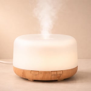 Può includere: Un diffusore di oli essenziali bianco e color legno con vapore che sale dalla parte superiore. Il diffusore ha una forma arrotondata con una parte superiore illuminata e una base con venature del legno. Il dispositivo è collegato con un cavo bianco.